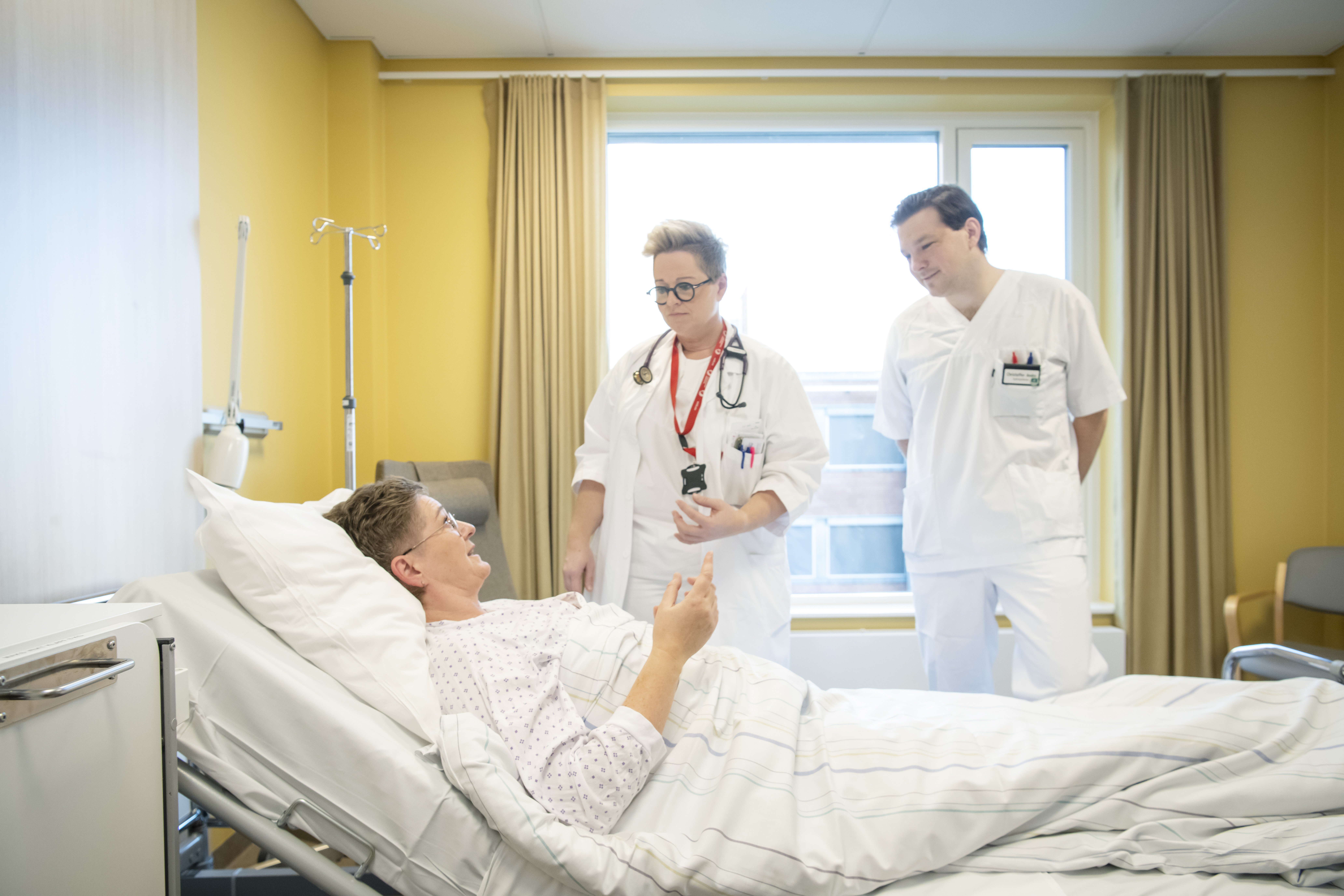 Kvinne ligger i sykehusseng. To leger snakker med henne.