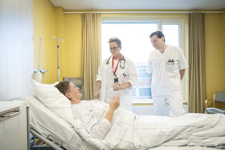 Kvinne ligger i sykehusseng. To leger snakker med henne.