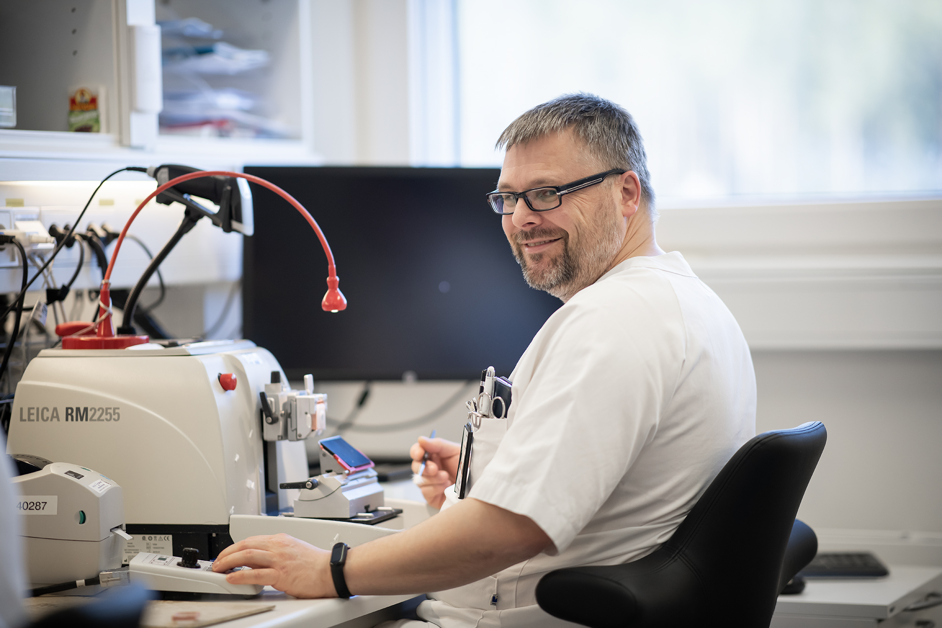 Mann smiler mot kamera, skjermer og mikroskop i bakgrunnen