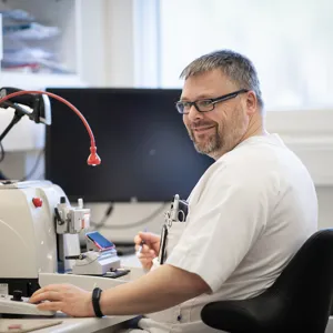 Mann smiler mot kamera, skjermer og mikroskop i bakgrunnen