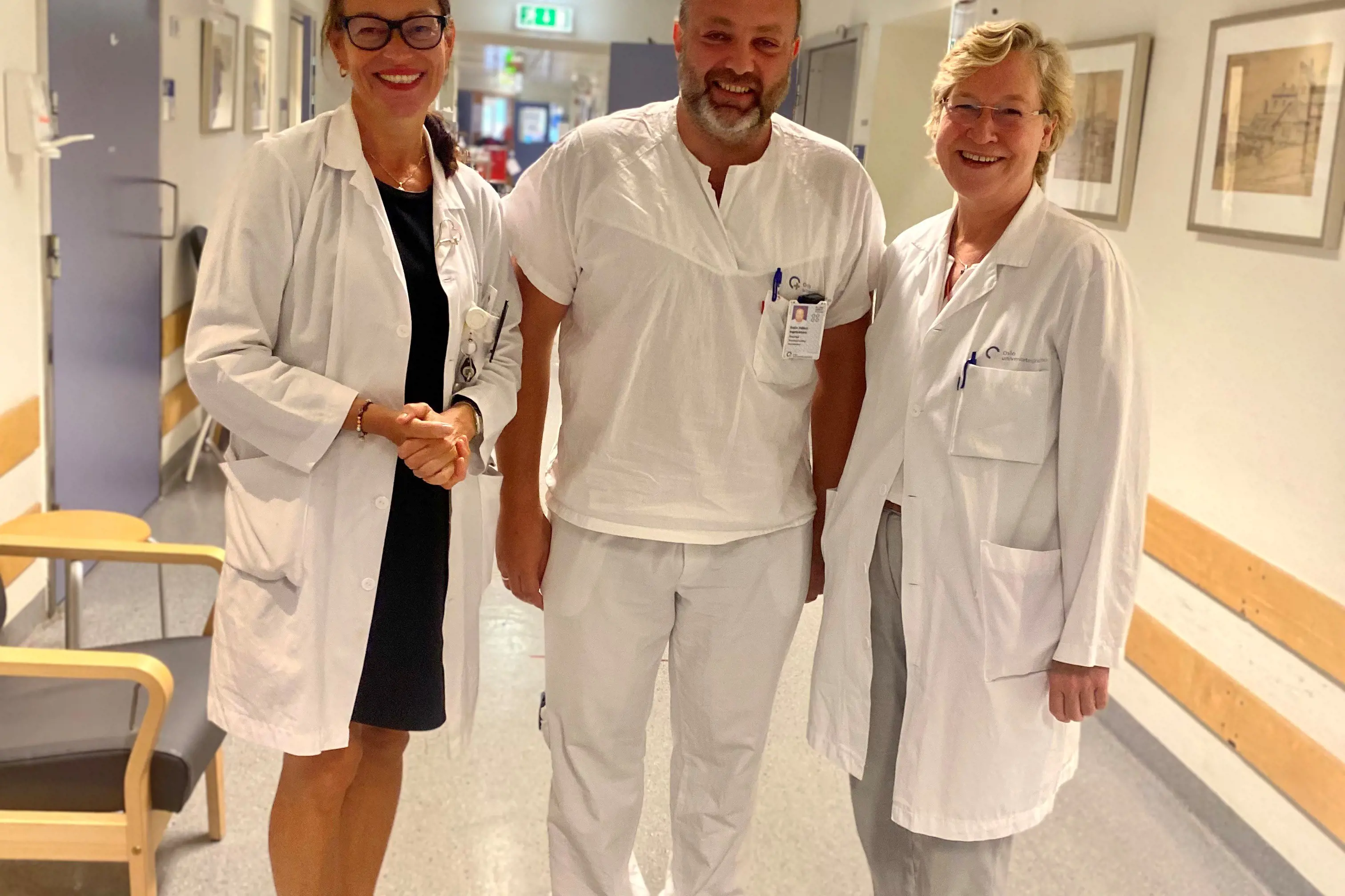 Tre smilende ansatte med hvite frakker i sykehuskorridor