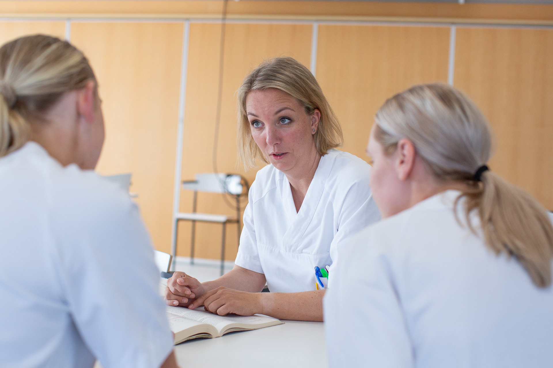 En kvinne i hvit skjorte, sitter ved et bord og samtaler med to andre.