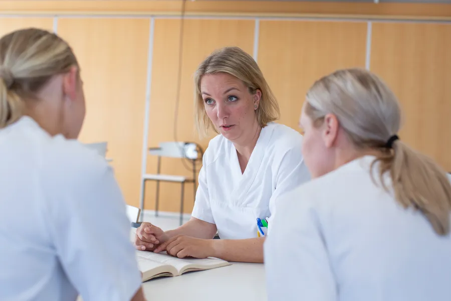 En kvinne i hvit skjorte, sitter ved et bord og samtaler med to andre.