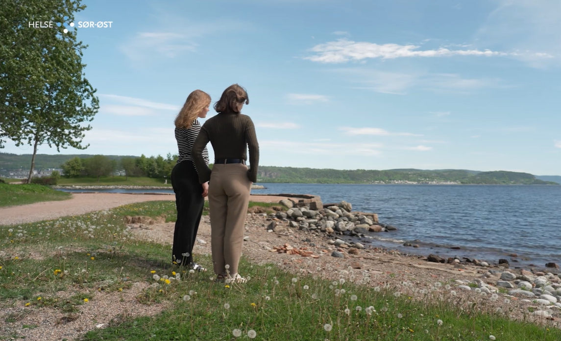 To kvinner som står med ryggen til på gresset ved sjøen.