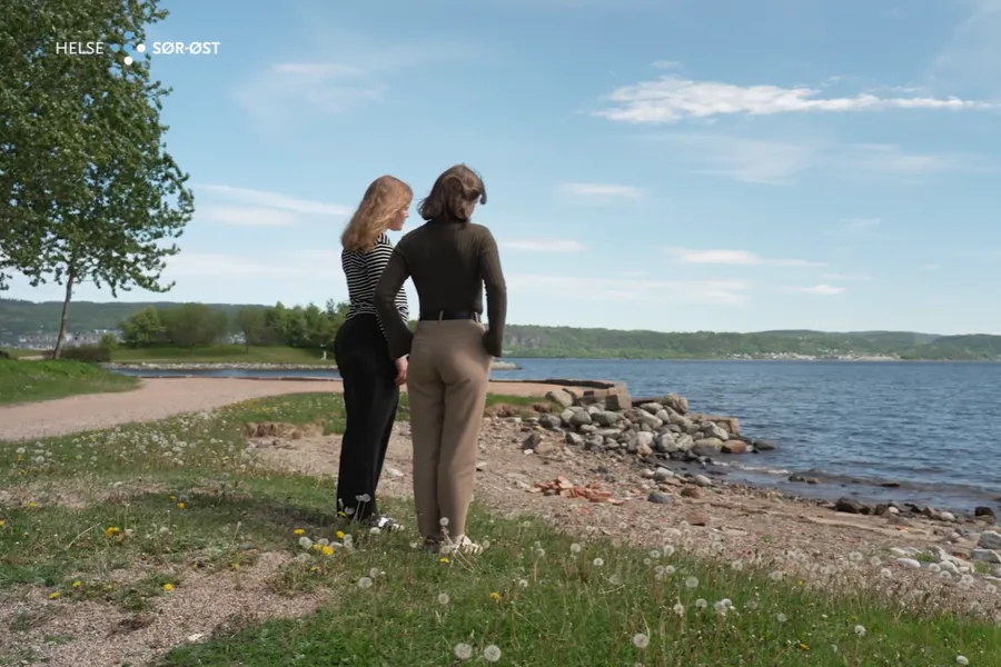To kvinner som står med ryggen til på gresset ved sjøen.