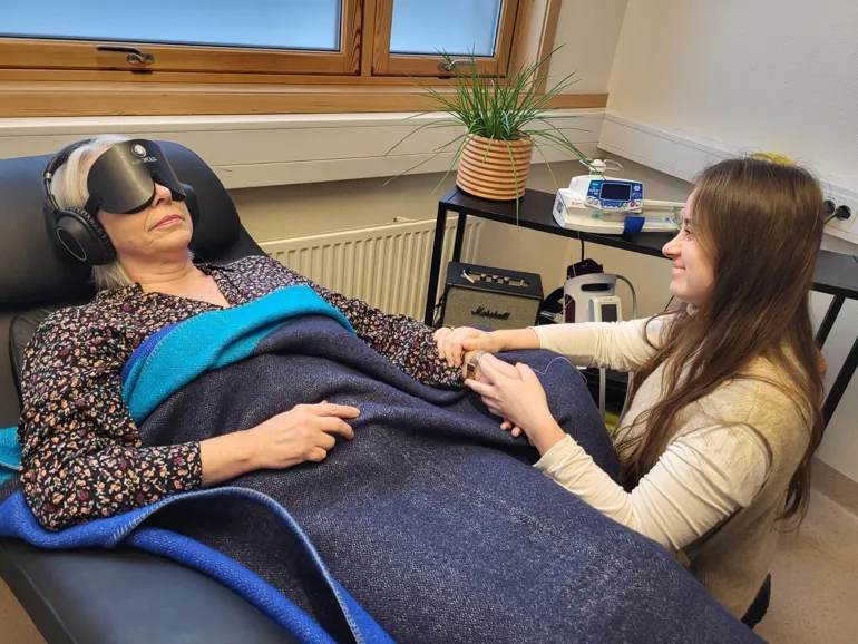 En kvinne ligger med hodetelefoner, sovemaske og teppe over seg i hvilestol, mens en annen kvinne holder henne i hånden.