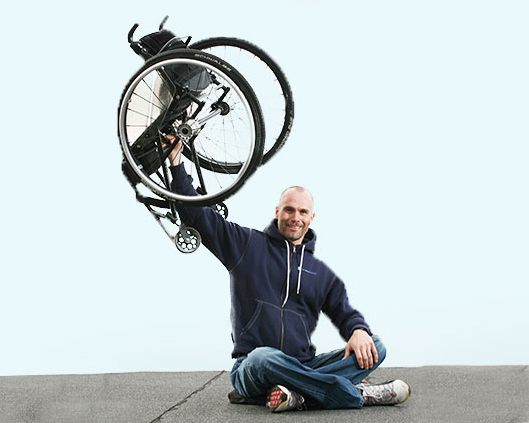 Mann sitter med bena i kors og holder en rullestol i en arm over hodet