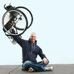 Mann sitter med bena i kors og holder en rullestol i en arm over hodet